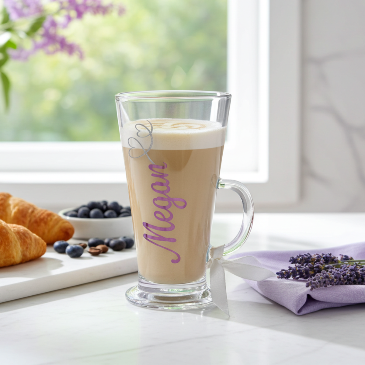 Personalised clear latte glass mug in a pink purple chrome holographic vinyl. Has the name 'Megan' with a frosted silver glitter heart swirl detail. Has a satin ribbon around its wide loop  handle. Glass filled with a latte drink styled on a white surface next to  berrries & lavender sprigs.
