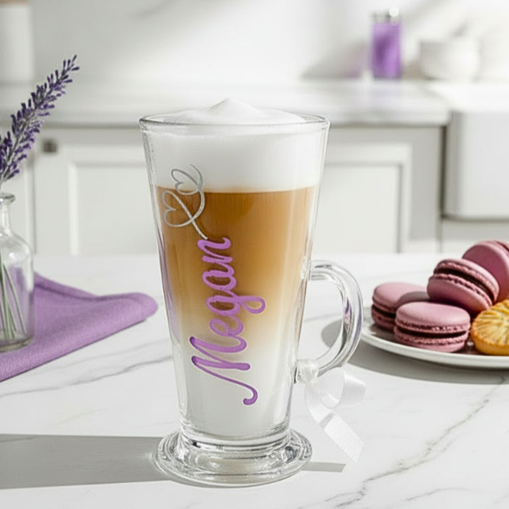 Personalised latte glass mug with the text 'Megan' on it in a holographic chrome pink purple vinyl. Features a silver glitter heart swirl design & a white satin ribbon on the handle. Placed on a marble counter top next to a plate of macaroons and a small vase of flowers.