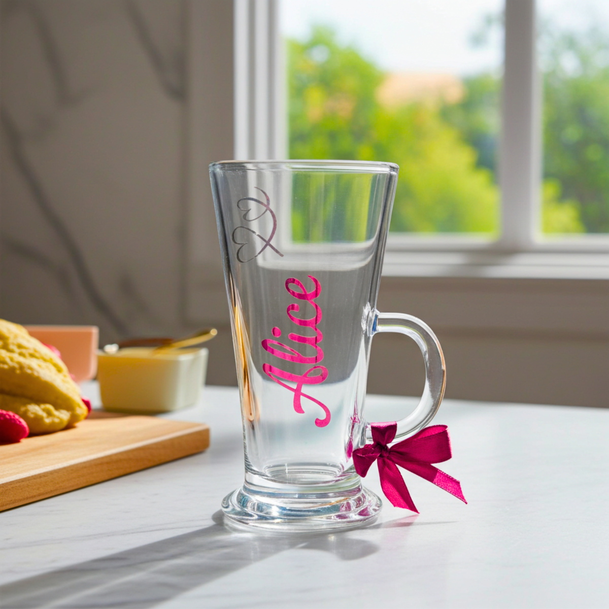 Personalised clear latte & hot chocolate glass with the text 'Alice' in a royal pink chrome holographic vinyl. This tapered coffee glass mug features a silver glitter hearts design & a royal pink satin ribbon tied around its wide loop handle. Placed on a white counter top.