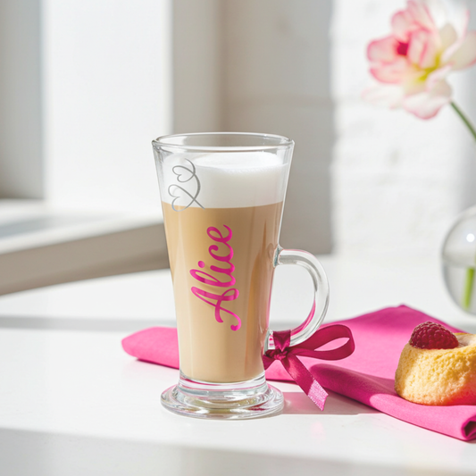Personalised latte glass filled with a latte drink with the text 'Alice' in a royal pink chrome holographic vinyl. This tapered coffee glass mug has a silver glitter hearts swirl design & a royal pink satin ribbon tied around its wide loop handle. Placed on a white surface next to a pink napkin & a pastry.