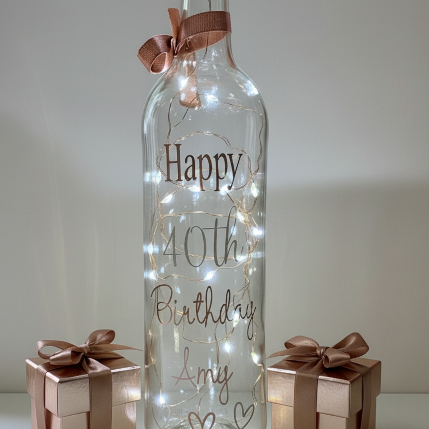 Personalised LED light up glass  bottle with 'Happy 40th Birthday Amy' in rose gold & silver vinyl text with small gift boxes each side.