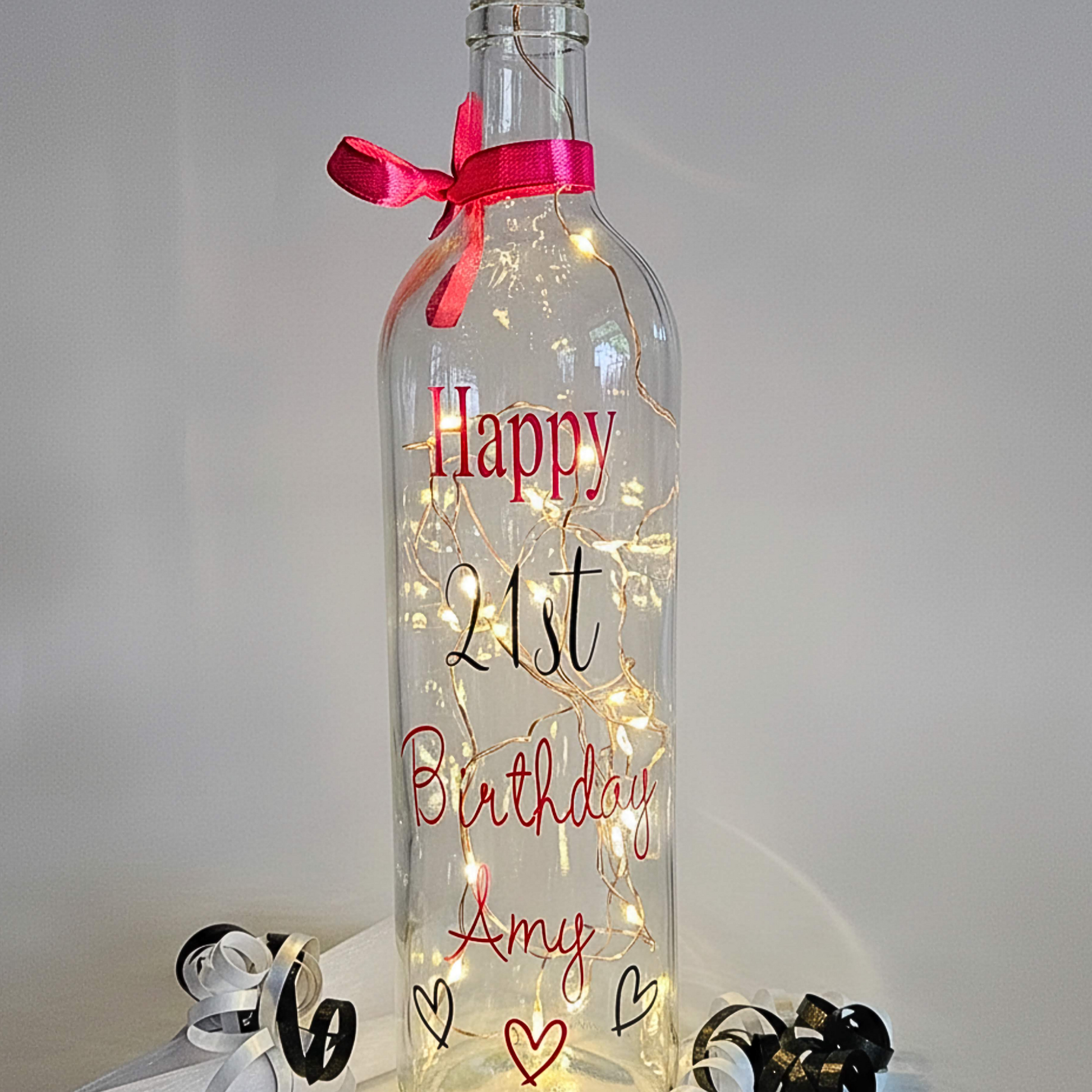 Personalised LED light up glass bottle with 'Happy Birthday Amy' in a hot pink vinyl text & age '21st' in black vinyl text next to an organza gift bag & satin curling ribbons.