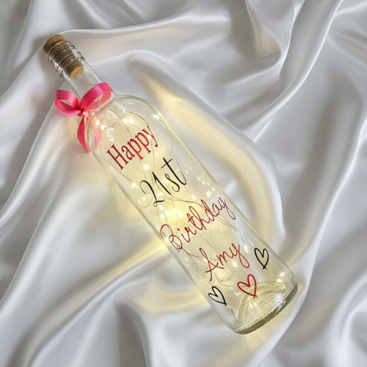Personalised 21st birthday LED light up bottle with the text 'Happy Birthday Amy' in a hot pink vinyl & age '21' in a glossy black vinyl. Features a pink and black hearts design and a hot pink satin ribbon around the bottle neck. Has warm white LED lights inside. Laid on a white satin drape.