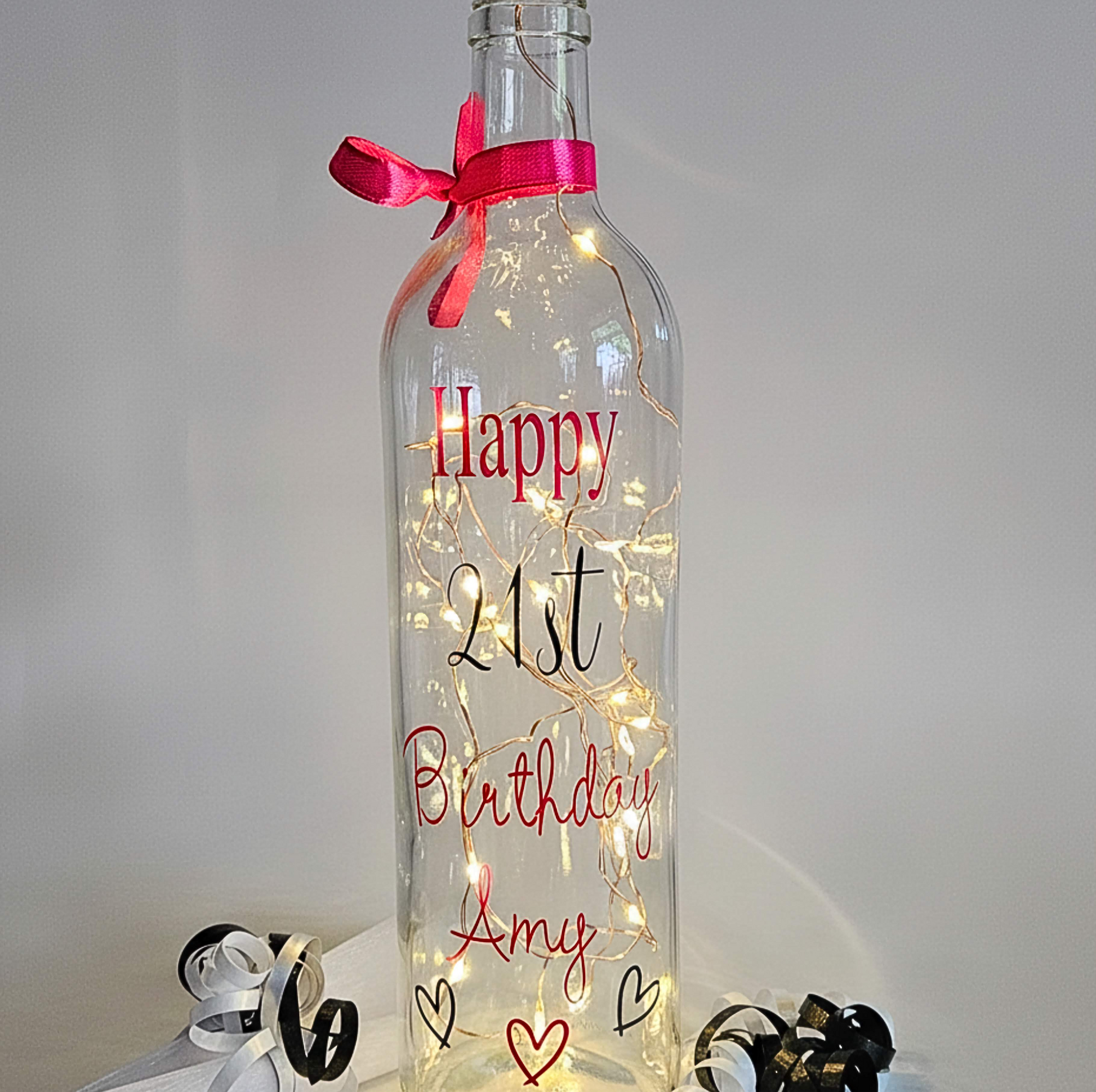 A clear bottle with hearts & lights. Has the wording 'Happy Birthday Amy' in pink and 21st in black. Has a ribbon around the neck & around
