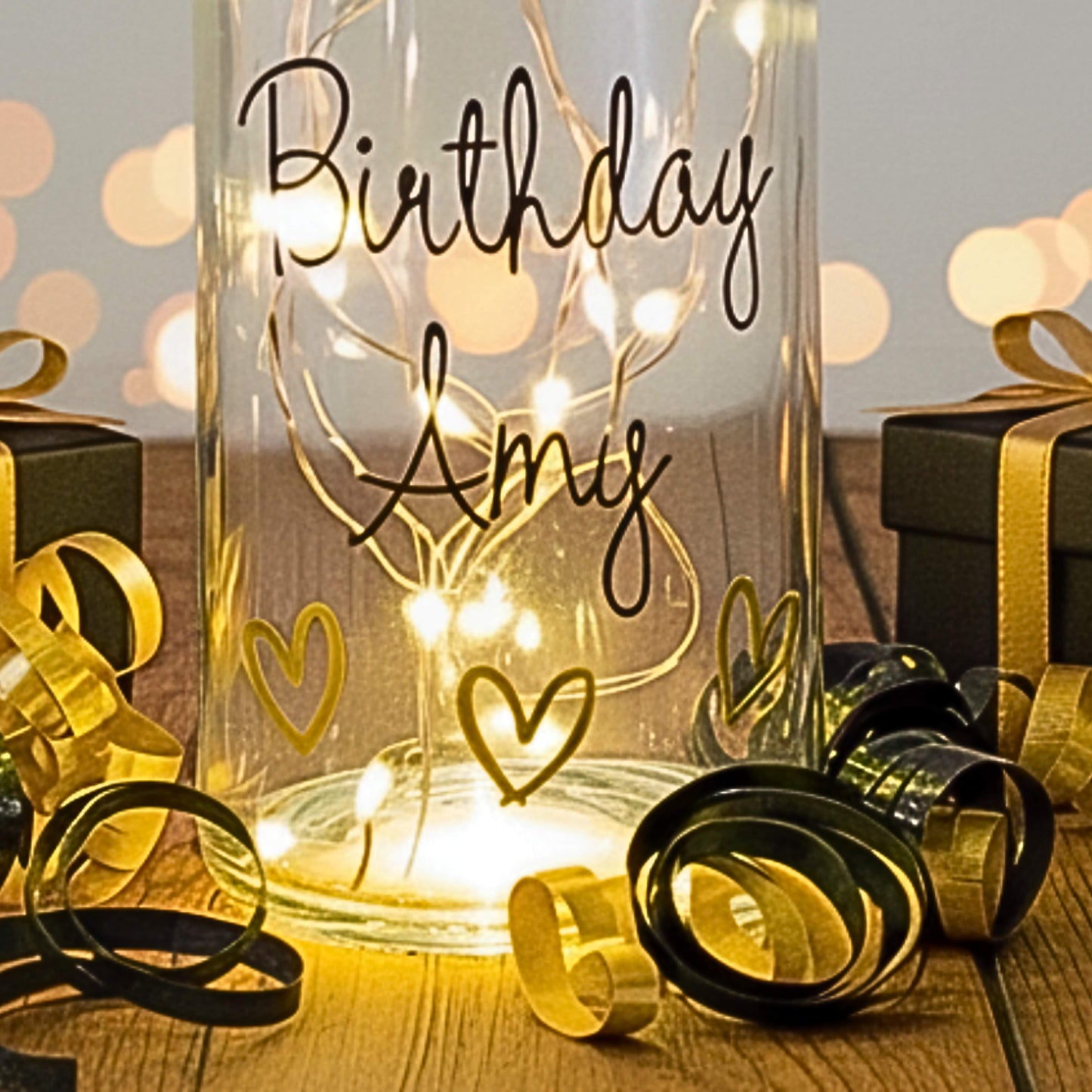 A clear glass bottle with led lights & the words 'Birthday Amy' with gold vinyl hearts. Has a background of gift boxes and ribbons