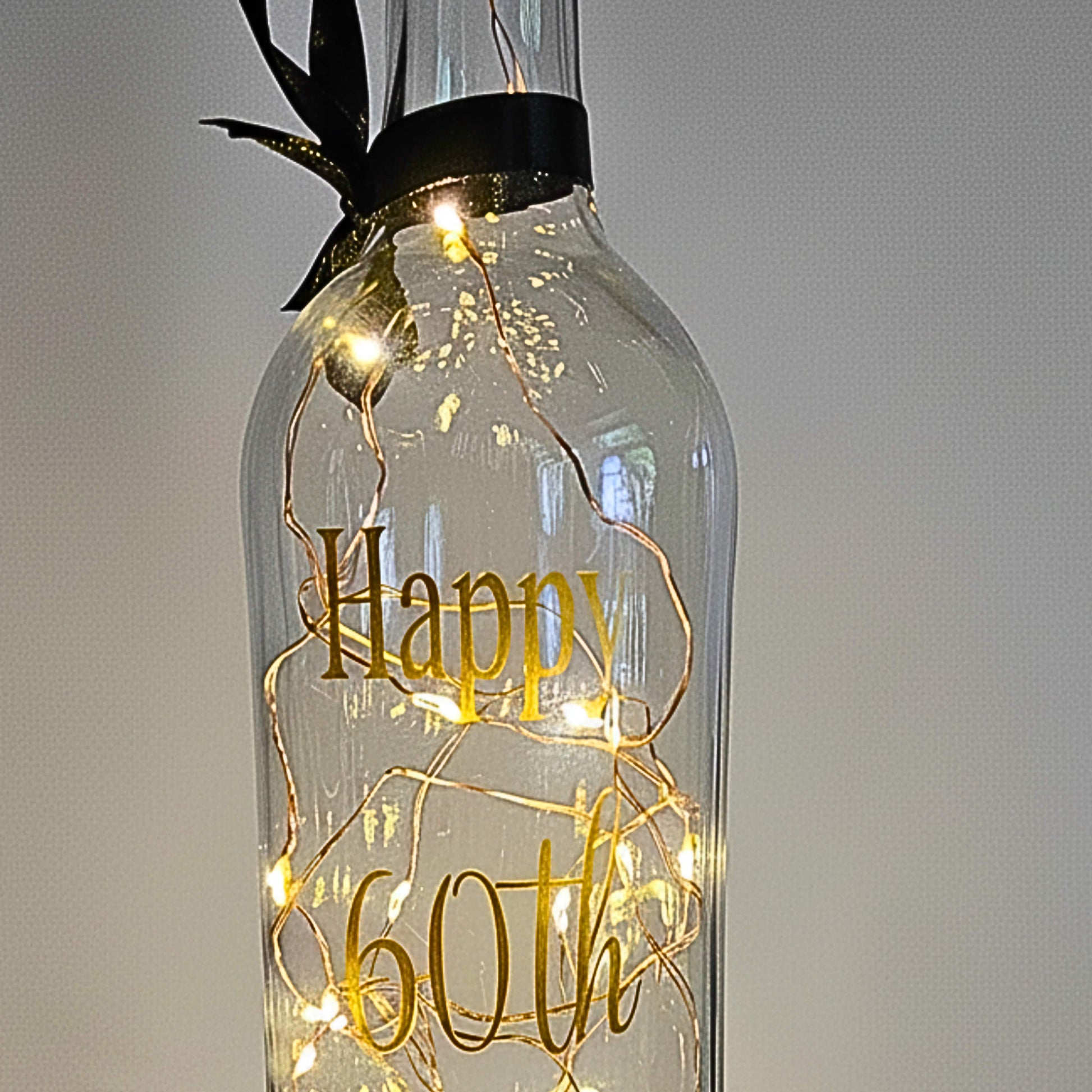 A clear bottle with lights. Has 'Happy 60th' in gold vinyl with a black ribbon around the neck.