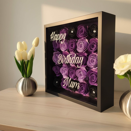 Personalised shadow box with the text 'Happy Birthday Mum' on it with a display of purple & black flowers behind a glass front. Sits on a shelf next to vase of flowers
