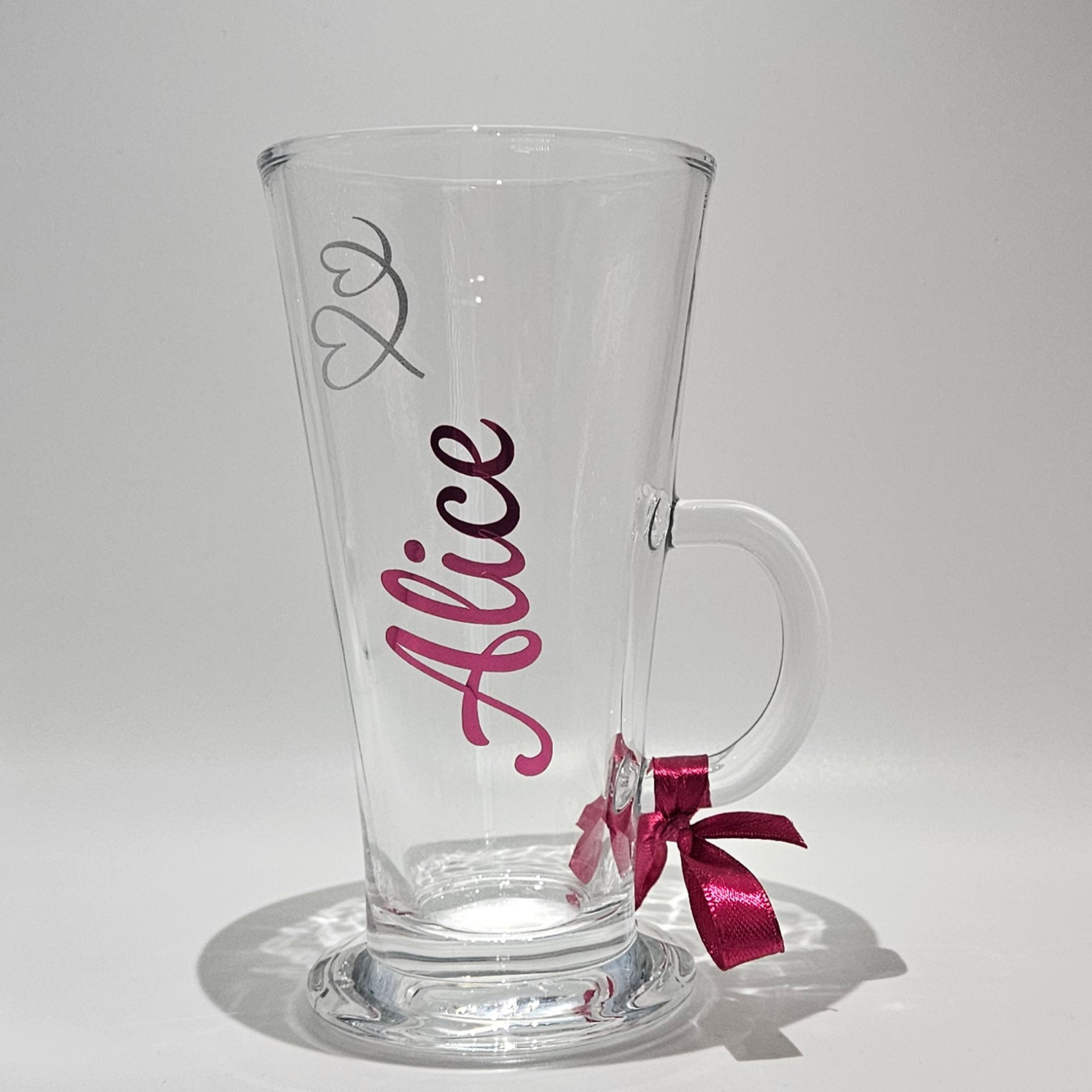 Personalised clear latte glass mug with the name 'Alice'on it in pink holographic vinyl. Has a pink satin ribbon on the handle on a white background.