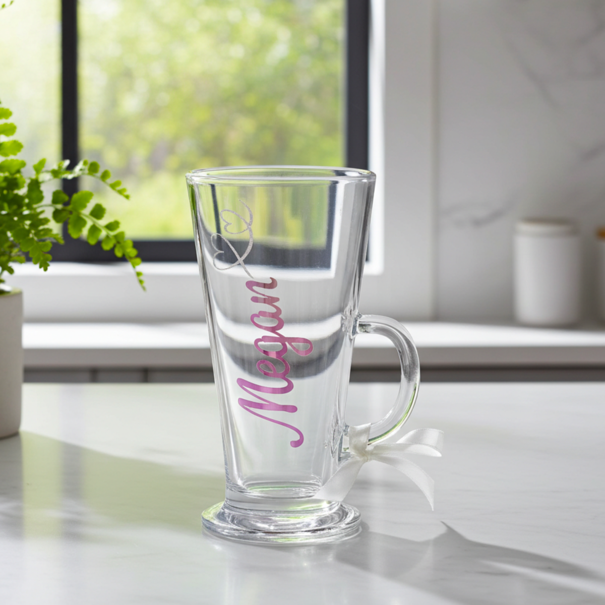 Personalised clear latte & hot chocolate glass with the text 'Megan' in a pink purple chrome holographic vinyl. This tapered coffee glass mug features a silver glitter hearts swirl design and a white satin ribbon tied around its wide loop handle. Placed on a white counter top in a kitchen setting.
