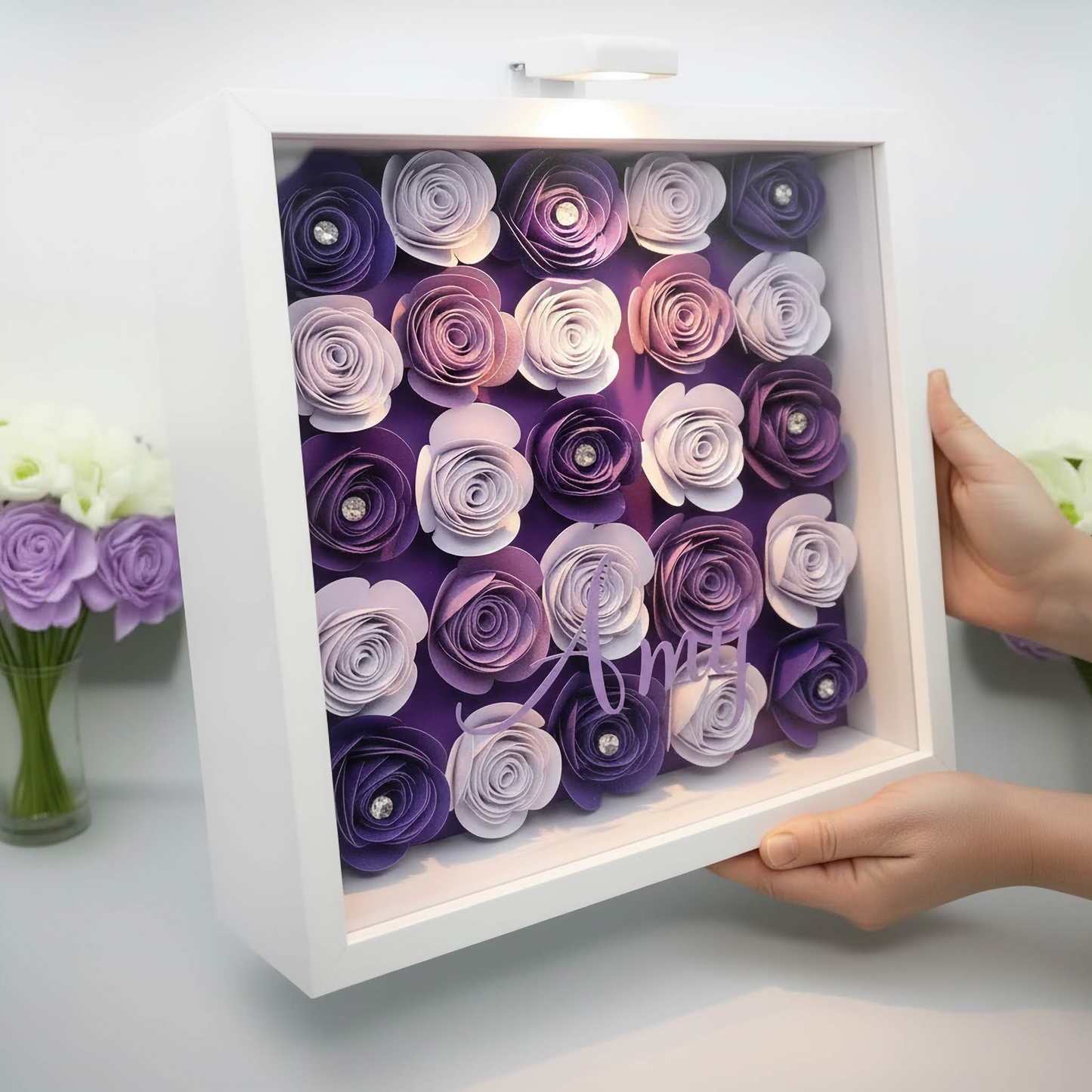Personalised 3D shadow box with the name 'Amy' on a glass front. Has a purple, lilac & white paper cardstock floral display in a white square frame held up next to a vase of flowers.