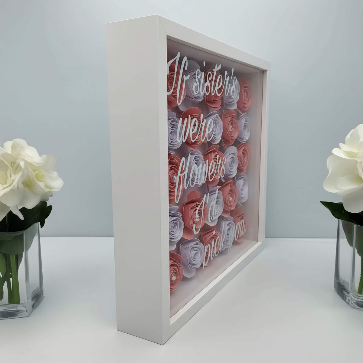 Personalised 3D shadow box with hand-rolled pink & white paper cardstock flowers in a white square frame and text on a glass front. Stood  in between two vases with white roses on a light background.