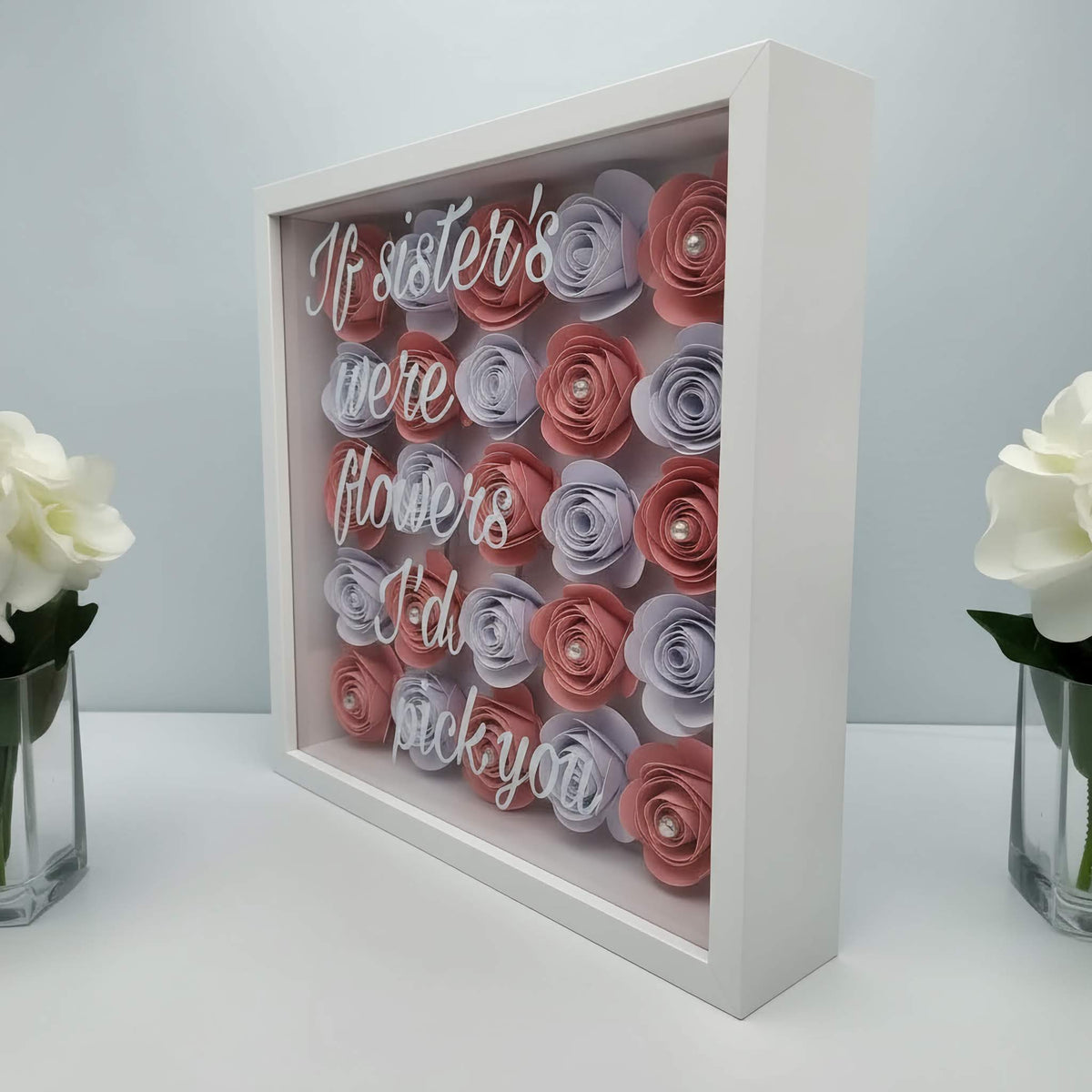 Personalised 3D shadow box with hand-rolled pink & white paper cardstock flowers in a white square frame and text on a glass front. Stood on a white surface with vases of white roses each side.