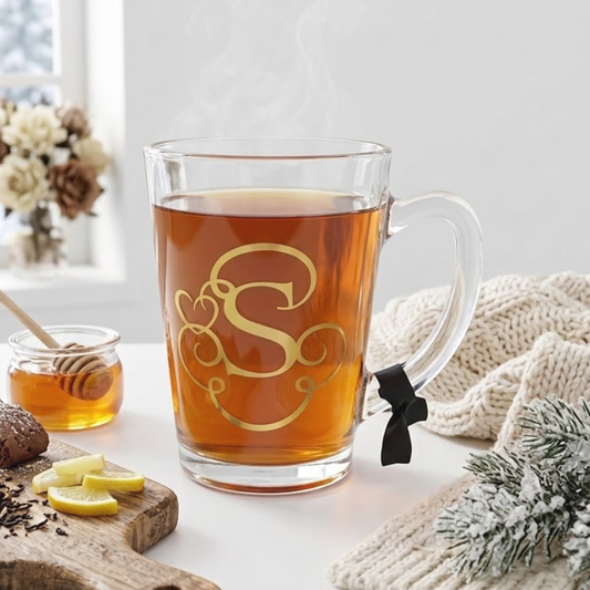 Personalised clear glass teacup mug filled with tea. Features a gold chrome vinyl monogram heart swirl design with the letter 'S'. Has a black satin ribbon tied around its wide loop handle. Placed on a white counter top on a plain background.
