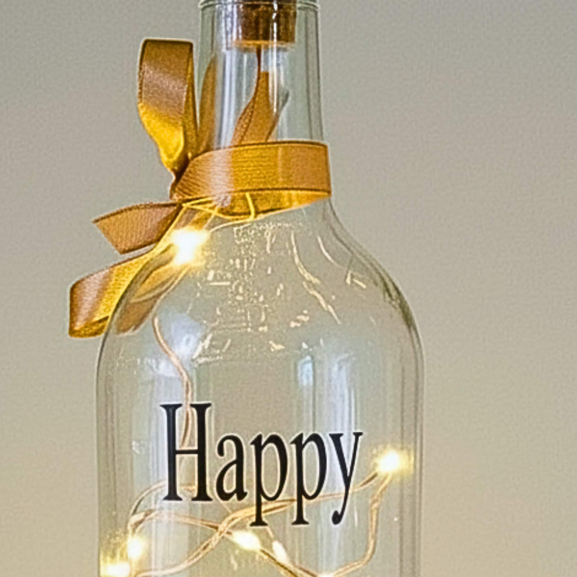 The top of a personalised LED light up glass bottle with the text 'Happy' & a gold satin ribbon on a neutral background