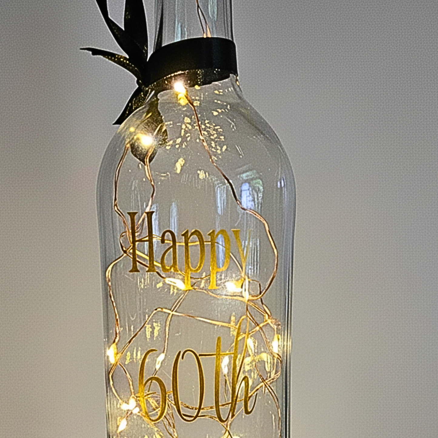 The top of a personalised LED light up glass bottle with gold text 'Happy 60th' on a plain background