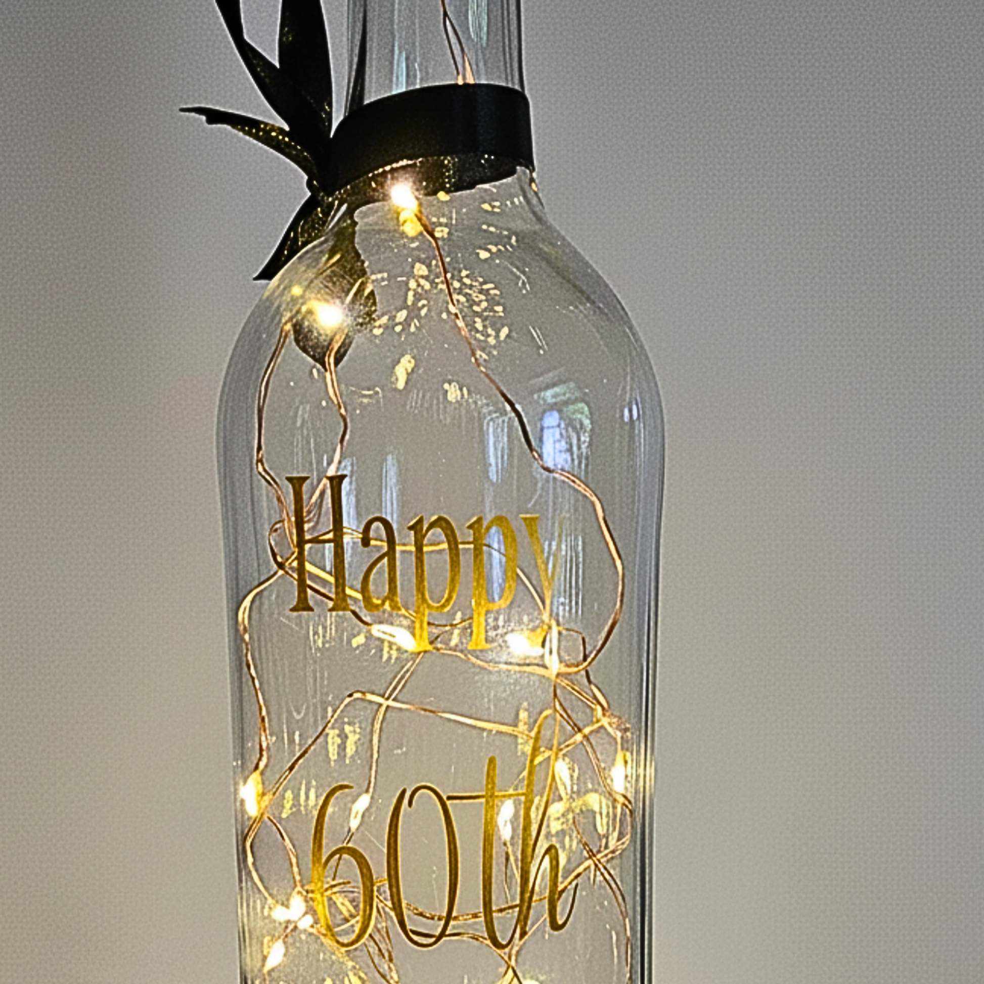 The top of a personalised LED light up glass bottle with gold text 'Happy 60th' on a plain background